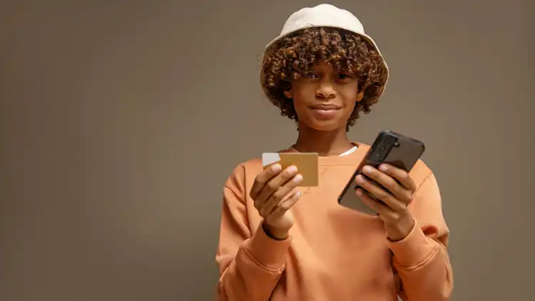 Boy stands with a credit card in one hand and his mobile in the other