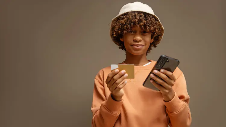 Boy stands with a credit card in one hand and his mobile in the other